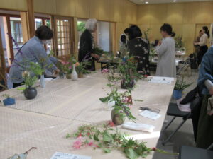 手前のテーブルには花が生けられた小ぶりの花瓶が複数置かれ、複数の参加者が奥のテーブルの周りに集まって説明を聞いている様子の写真