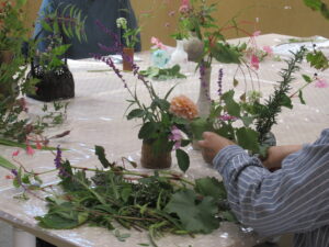 青いストライプのシャツを着た人物が、テーブルに広げられたダリアなどの色鮮やかな花材を手に取り、器へのバランスを考えながら丁寧に生け花をしている様子の写真