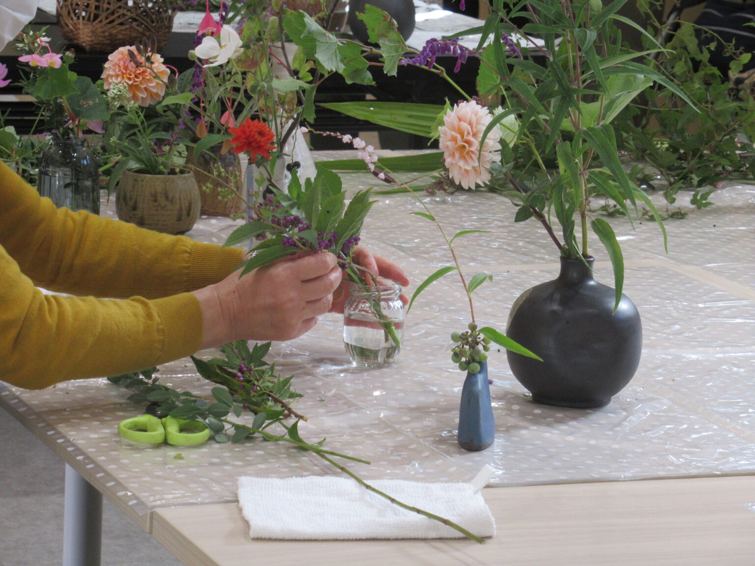 透明のシートを敷いたテーブルの上に、花を生けた様々な花瓶が複数置かれ、手前では透明な小瓶に紫色の小さな実をつけた枝を丁寧に生けている様子の写真