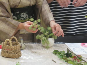 着物姿の女性が、白い丸みを帯びた小さな花瓶に薄緑色の小さな花が密集した植物を丁寧に生けており、手元には小さな編み籠や切り取られた葉が置かれている写真