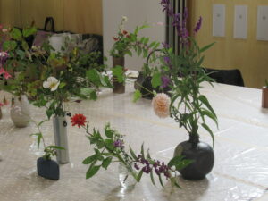 ビニールクロスの敷かれた大きなテーブルの上に、ダリアや野の花が活けられた大小様々な花瓶が並び、それぞれが異なる高さや広がりを持って配置されている様子を捉えた写真