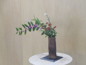 明るい室内にある丸い白木の台座の上に、小さな紫の実をたくさんつけた枝が左側に大きくしなり、右側には赤や白の小花が直立するように生けられた花瓶を写した写真