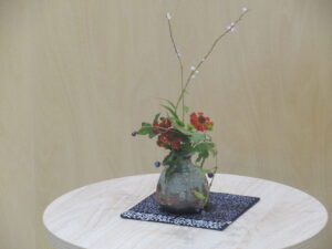 丸い白木の台の中央に、赤い小花や紫の実、そして高く伸びた白い蕾を持つ枝がバランスよく生けられた味わい深い陶器の花瓶が置かれた写真