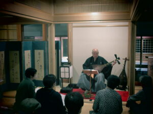 静寂な和室で赤い毛氈の上に座る奏者が薩摩琵琶を演奏し、その前で数人の観客が静かに聴き入っている写真