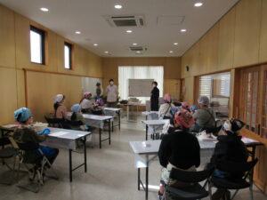 明るい木目調の壁の室内で、三角巾を被ったたくさんの親子の参加者たちが長机に分かれて座り、前方のホワイトボードの前に立つ安藤先生の話を聞いている様子の写真