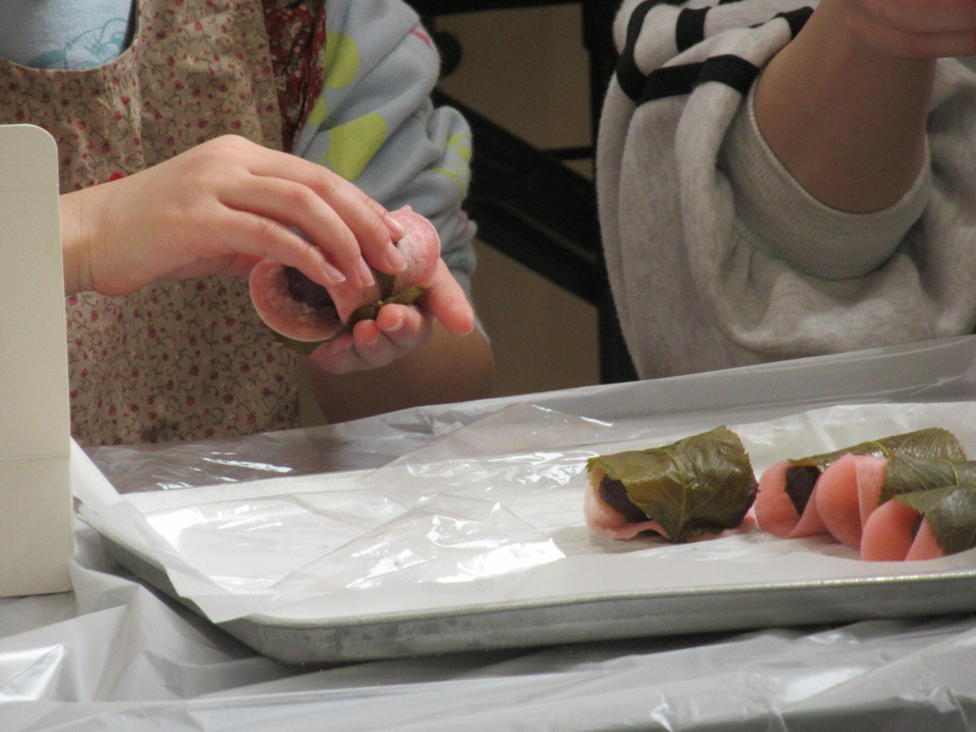 あんこをくるんだ薄いピンク色の生地に塩漬けの桜の葉を丁寧に巻き付けている手元と、出来上がった桜餅が作業台の上のバットに並んでいる様子の写真