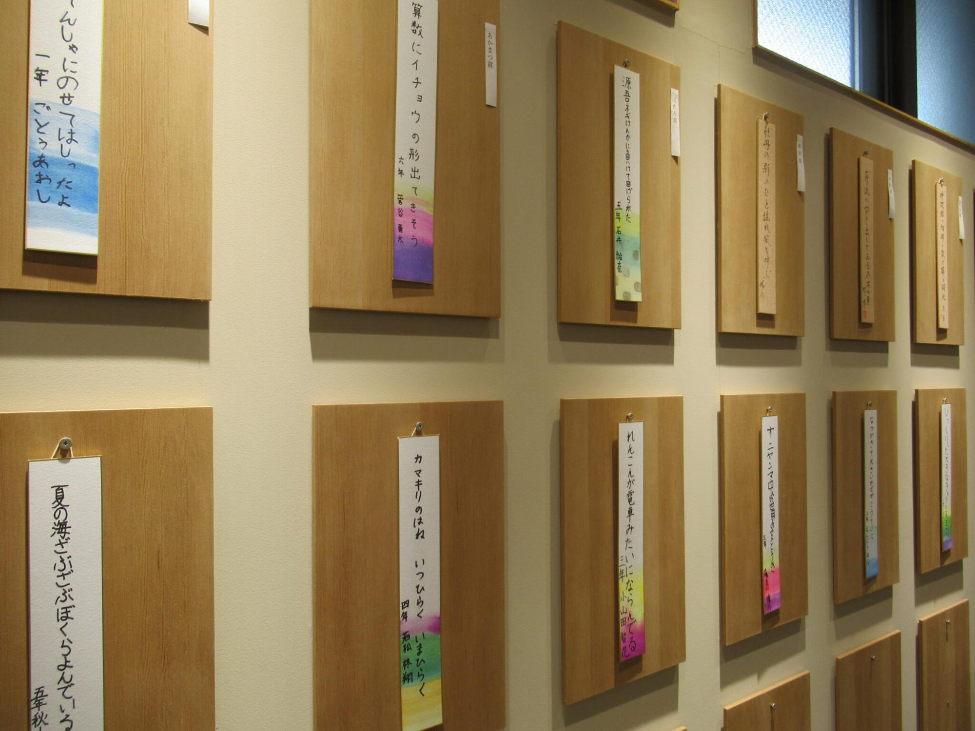 壁面に整然と並べられた木製ボードに、色鮮やかな水彩風の装飾が施された短冊や茶褐色の短冊が掲示され、子どもたちの手書きによる俳句作品が一つずつ丁寧に展示されている展示会場の風景を斜めから捉えた写真