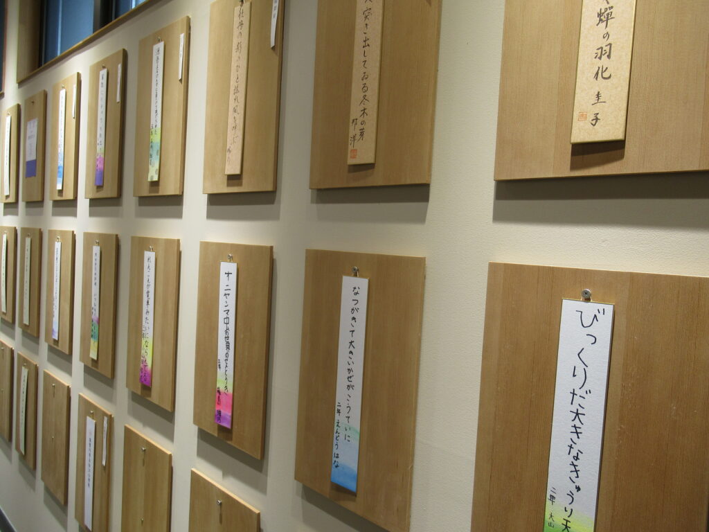 壁面に整然と並べられた木製ボードに、色鮮やかな水彩風の装飾が施された短冊や茶褐色の短冊が掲示され、子どもたちの感性が光る俳句作品が一つずつ丁寧に展示されている写真