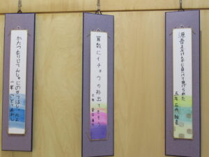 木目調の壁を背に、色鮮やかな水彩画の短冊が吊るされ、小学校低学年から高学年の児童による俳句が3枚展示されている写真