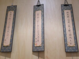 木目調の壁を背景に、和柄の縁取りが施された縦長の展示台紙が3枚並び、俳句が書かれた短冊が丁寧に固定され展示されている写真
