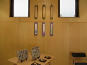 木目調の壁を背に、上段には落ち着いた茶系の短冊が3枚、下段には華やかな装飾が施された色の台紙付き短冊が3枚、計6枚の俳句短冊が整然と吊るされ、手前の机には受賞作品を刻んだ石板や記念品が並べられた写真