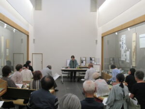 両側にガラスの展示ケースがある展示室で、前方に立つ講師の鈴木邦子さんと、座って話を聞いている多くの参加者の様子を後方から撮影した写真