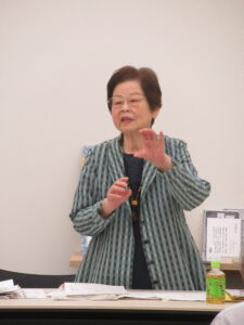 講師の鈴木邦子さんが資料を並べたテーブルの前に立ち、手振りを加えながら話をしている様子の写真
