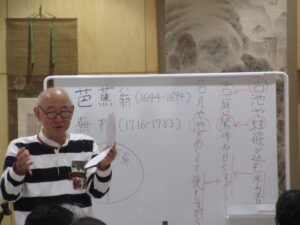 ホワイトボードの前に立つ講師が、松尾芭蕉の有名な句「古池や蛙飛びこむ水の音」などの解説を熱心に行い、芭蕉と蕪村の生没年や俳句の構造が板書された、文化講演会における学習の様子を捉えた写真