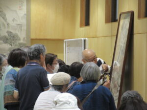 明るい木目調の壁に囲まれた室内で、講師が壁に立てかけられた大きな掛け軸を指し示しながら解説を行い、その周りを大勢の受講生が熱心に覗き込むようにして集まっている、文化講演会での作品鑑賞の様子を捉えた写真