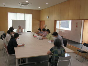 明るい会議室で数人が長机を囲んでメモを取りながらホワイトボードの前の席に座った講師の説明を聞いている様子の写真