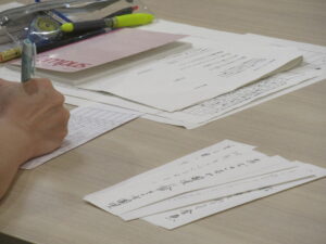 資料やノートを置いた机の脇に俳句が書かれた短冊が重なるように置かれ、その横で用紙に記入をしている人物の手元の写真