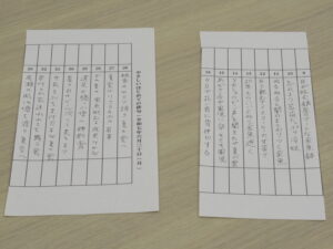 机上に縦の区分欄が並ぶ二枚の手書き表が横に並べて置かれ、細かい文字がびっしり記入された様子の写真