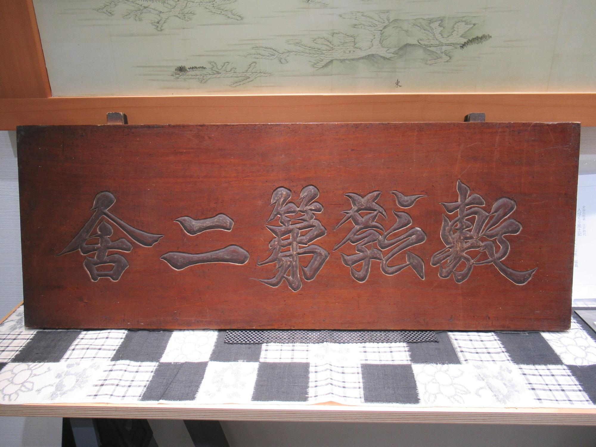 市松模様の布の上に置かれた経年変化を感じさせる重厚な木板の表面に「敷教第二舎」という文字が右から左へ一文字ずつ力強く彫り込まれている看板を正面から捉えた写真