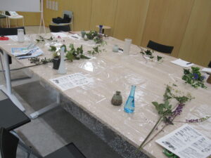 透明シートを敷いた大きなテーブルの上に、花材や花瓶などの花器、資料が置かれ、準備が整えられた作業台の様子の写真