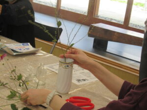 参加者が白い花器に細い枝物を差し込みながら配置を整え、手元には花材やハサミが置かれている作業中の写真