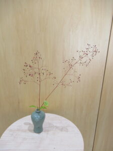 白木の丸い台の上に、赤い小さな実をつけた細枝と緑の小さな葉を生けた、水色の花瓶が置かれた写真