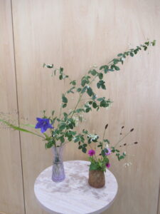 白木の丸い台に、紫の花と緑の葉をつけた長い枝ものなどが生けられたガラスの花瓶と、濃淡の桃色の花と細長い葉などが生けられた茶色の花瓶が並べられた写真