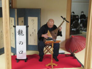 畳の上に敷かれた赤い毛氈の上に「龍の口」と書かれた題目が置かれ、その横の椅子に座った山田寛水氏が薩摩琵琶を弾いている様子の写真