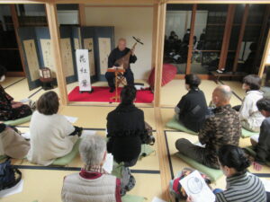 和室の柱で囲まれ赤い絨毯が敷かれたスペースに設置された椅子に薩摩琵琶を抱えた山田寛水氏が座り、その前にたくさんの参加者が座布団を敷いて座っている、催しの様子全体の写真