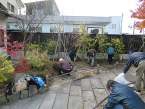 石畳の通路が続く日本庭園のような屋外で、数人の人々が腰をかがめ竹箒などを使って、落ち葉拾いや草むしりなどの清掃作業に励んでいる写真