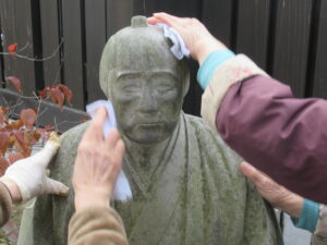 和装姿の相楽等躬を模した石像の頭部や肩を、数人の人々が白い布を使って丁寧に拭き掃除している様子を間近で捉えた写真