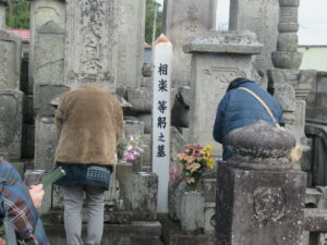 歴史を感じさせる数多くの古い石碑が並ぶ墓地の一角で、中央に立つ「相楽等躬之墓」と記された白い標柱に向かって二人の人物が深く頭を下げて手を合わせ、色鮮やかな供花が添えられている静かなお参りの風景を捉えた写真