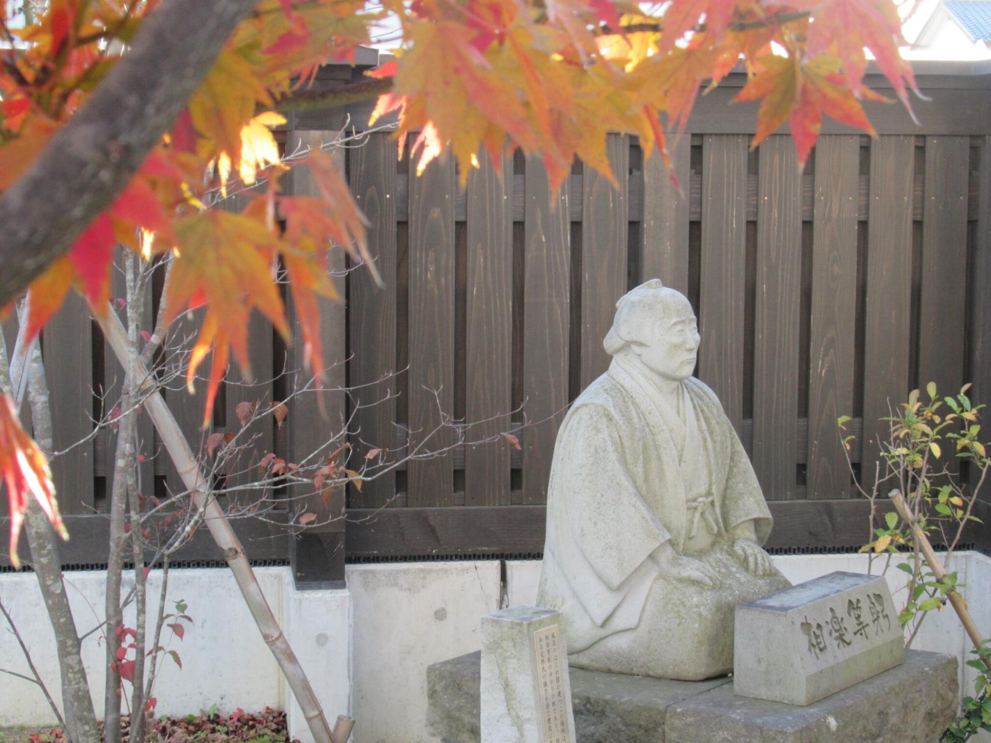 紅葉したカエデの葉が手前に広がり、奥には木造の柵を背景に和装姿で正座する相楽等躬の石像が置かれ、足元には「相楽等躬」と刻まれた石碑が並んでいる、秋の静かな庭園の風景写真
