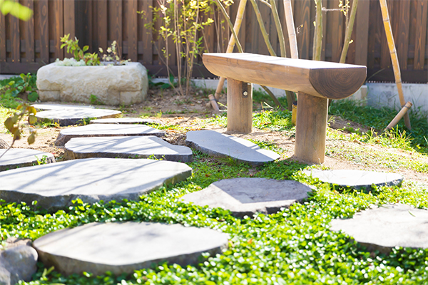 柔らかな日差しが差し込む庭に不規則な形の飛び石が連なり、その奥に木製のベンチが置かれた緑豊かな庭園の写真