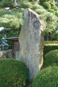 庭園の緑に囲まれた苔むした縦長の石碑が立ち淡い刻字が陽光に浮かび上がる静かな情景の写真