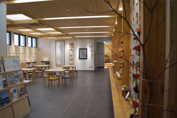 木材を多く用いた明るい室内に複数の机と椅子が置かれ、壁面には書画や飾りが展示された落ち着いた雰囲気のオープンギャラリーの写真