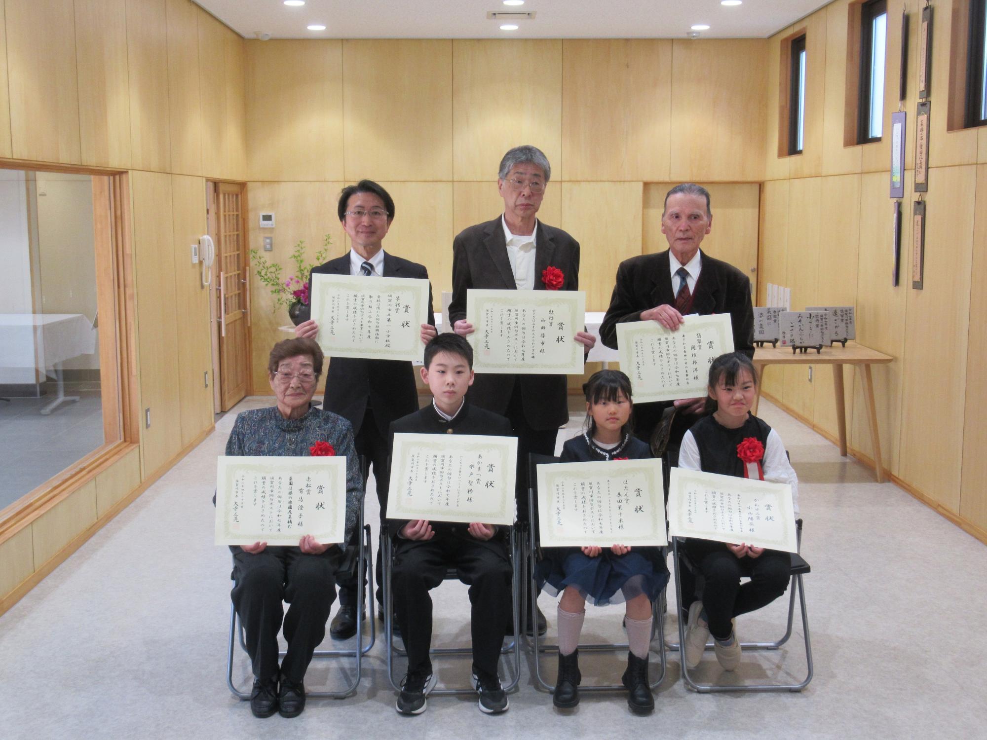 木目調の明るい室内で、表彰状を手にした7名が、前列と後列に分かれて正面を向き、晴れやかな表情の集合写真