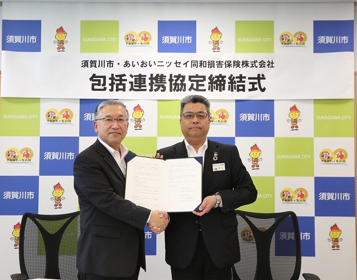 須賀川市とあいおいニッセイ同和損害保険株式会社の「包括連携協定締結式」の文字が書かれた看板の前で、スーツ姿の二人の男性が書類を挟んで握手をしている様子を写した写真