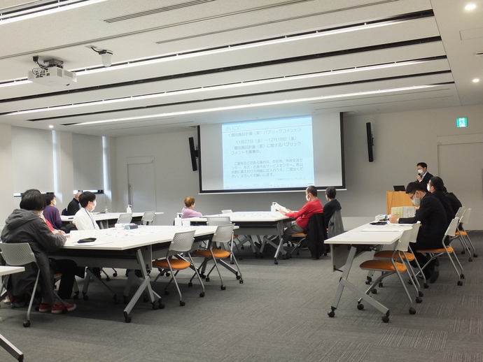 会議室に複数並んだ白いテーブルに複数のが座り、右側に関係者が座る中、前方のスクリーンに資料を写しながら、演台に立った男性がマイクで話をしている様子の写真