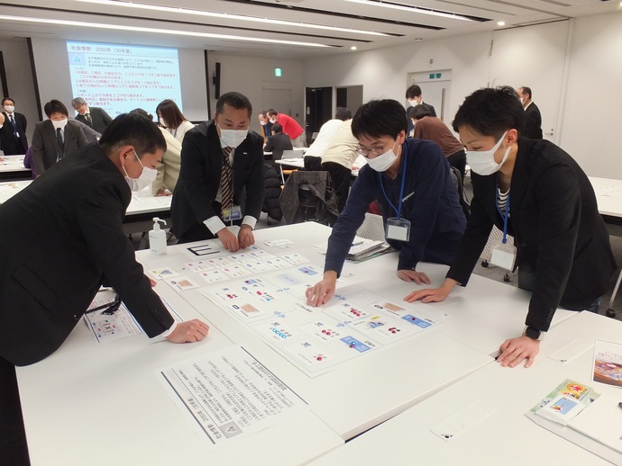 会議室の前方のスクリーンに資料が映し出され、複数のグループに分かれた参加者たちが、それぞれ資料を広げたテーブルを囲んで話し合いをしている様子の写真