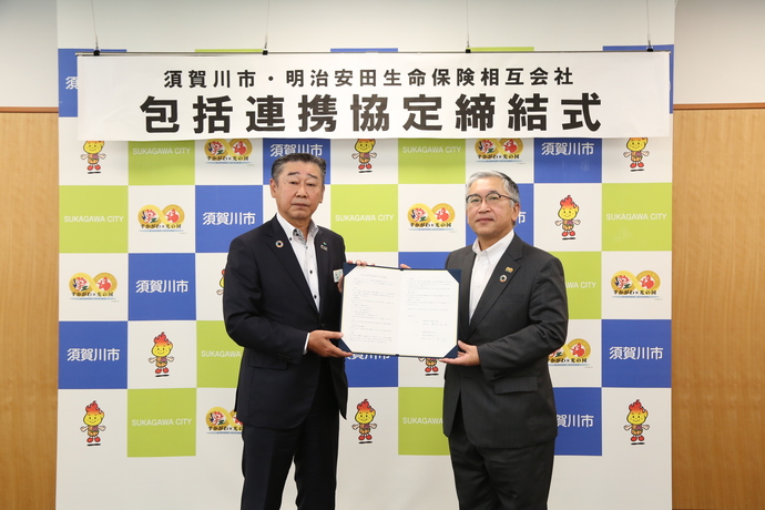 須賀川市明治安田生命保険相互会社包括連携協定締結式と書かれた横断幕を背景に橋本市長と大杉郡山支社長が協定書を一緒に持ち記念撮影を行っている様子の写真