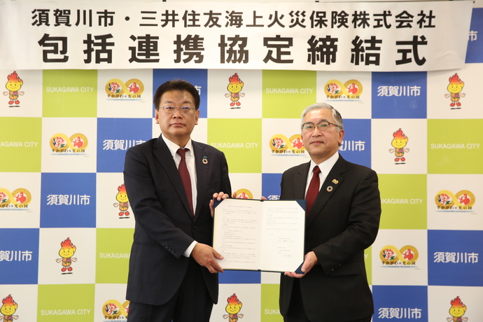 包括連携協定締結式と書かれた横断幕を背景に橋本市長と大島支店長が、締結書を一緒に持ち記念撮影を行っている写真