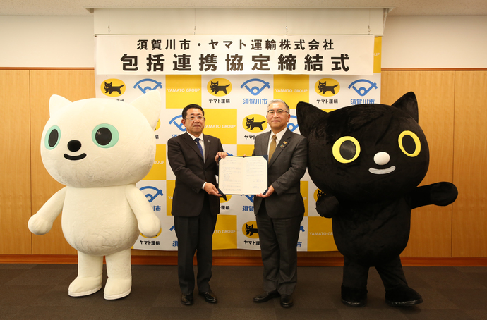 包括連携協定締結式と書かれた横断幕を背景に橋本市長と齊藤郡山主管支店長が締結書を一緒に持ちクロネコとシロネコの着ぐるみと一緒に記念撮影を行っている写真