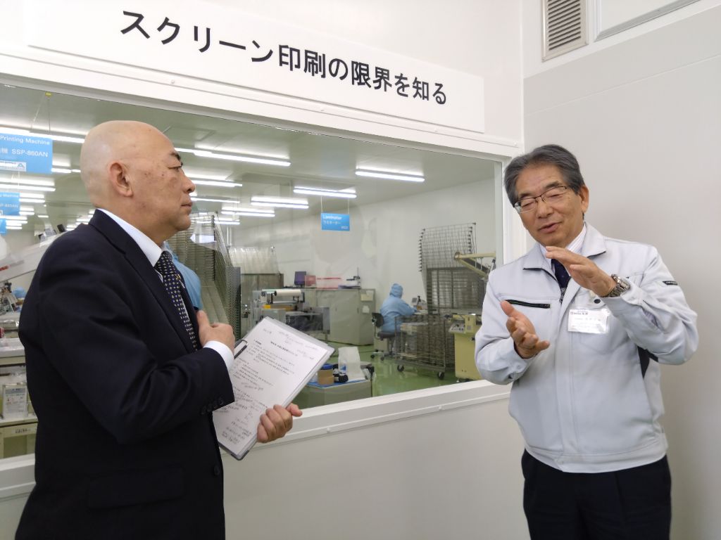 工場の組立ラインで複数の作業者が青や濃紺の作業着を着て製品を組み立てており、手前には緑色の棚と段ボール箱が置かれ、佐藤事業部長が液晶ユニット製造を男性に解説している様子の写真