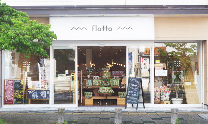 歩道沿いの白を基調とした清潔感のある店構えに「flatto」というロゴが大きく掲げられ、大きなガラス張りの入り口からは店内にたくさんの商品が並んだ様子が見えている、すかがわ観光物産館flattoの外観写真