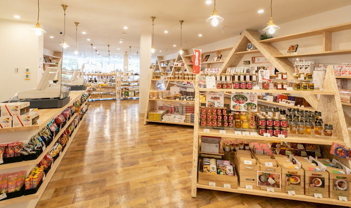 木目調の温かみのある床が広がる白い壁の店内に、瓶詰めや箱詰めなどの商品が美しく陳列された山型の木製棚が並び、天井から吊り下げられたライトが優しく空間を照らしている、すかがわ観光物産館flatto の内観写真