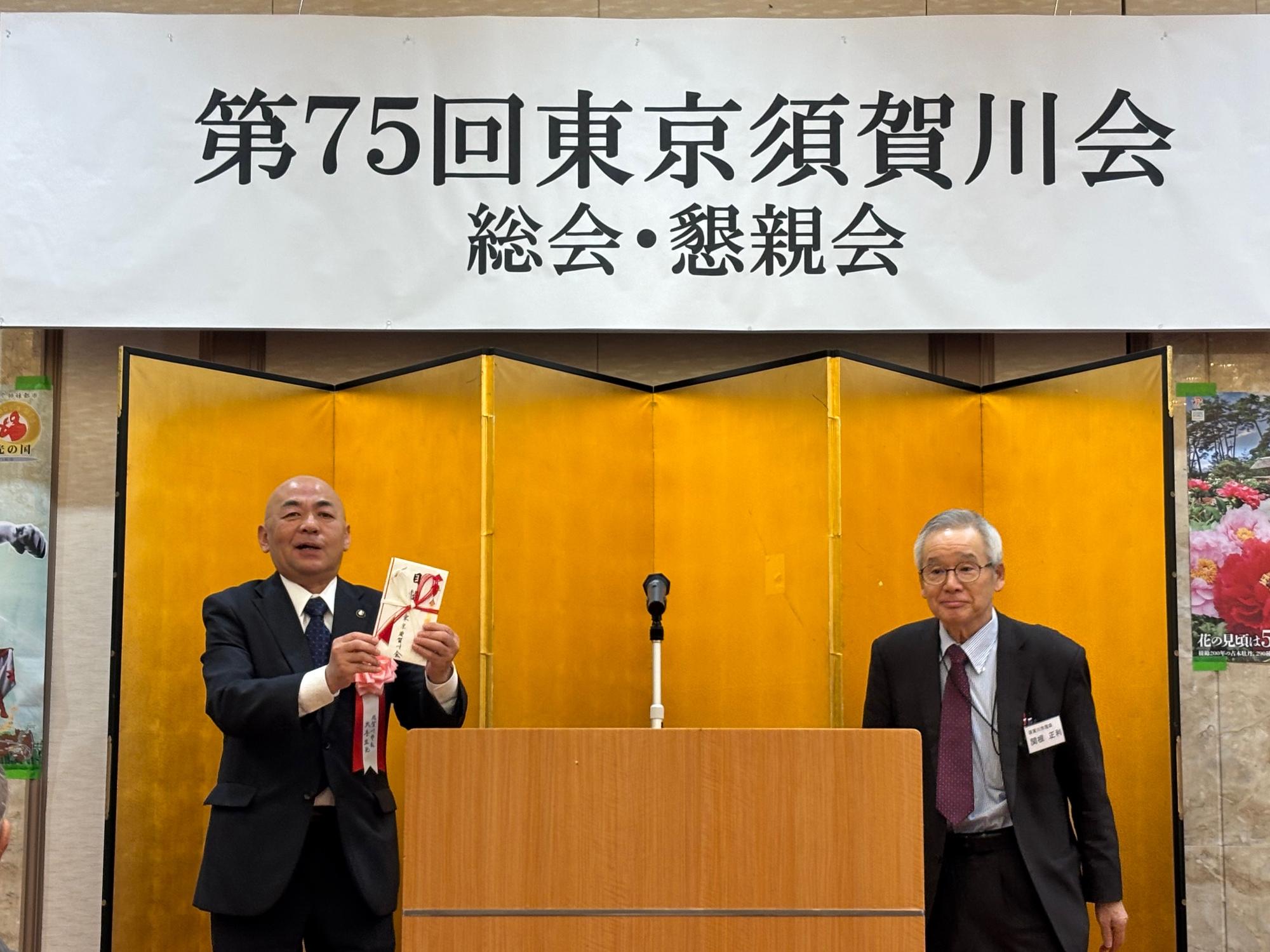 「第75回 東京須賀川会 総会・懇親会」の横断幕が掲げられ、金屏風が設置された演台に関根正利会長と大寺市長が並んで立っている様子の写真
