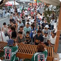 お祭りで賑わう通りの様子。手前では緑の法被を着た人が作業し、奥まで多くの参拝客や見物人が並んでいる。