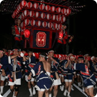 夜のお祭りで、紅白の提灯を何重にも積み上げた大きな山車を、大勢の担ぎ手たちが力強く引いて練り歩く光景。