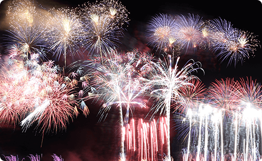 夜空いっぱいに打ち上がる、白や赤、紫色の華やかな花火。火花が何重にも重なり、夜空を明るく彩っている